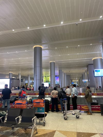 The image depicts a busy airport terminal, likely in Gollapalle Khurd, India, with people standing in a queue, possibly for check-in or security. The scene is indoors, with a modern architectural design featuring high ceilings, prominent pillars, and a tiled floor. A long line of passengers with luggage suggests an ongoing travel process.

Several individuals are visible, some with backpacks and trolleys, indicating they are travelers. The activity appears to be the typical flow of passengers at an airport. The lighting is bright and artificial, suggesting it could be any time of day.

Notable details include digital displays and advertisements mounted on pillars and walls, one of which shows the text "Your FEEDBACK" and what appears to be a feedback form. Another display shows "60". The visible text also includes "punjab national bank pnb" on signage, suggesting the presence of banking services or advertisements within the terminal. The overall atmosphere seems to be one of transit and routine airport operations. The presence of luggage trolleys and various bags confirms the setting as a travel hub.