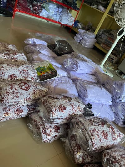 The image depicts a storage area, likely a retail space or warehouse, filled with packaged garments. Stacks of white and light purple folded clothing, individually wrapped in plastic, are piled on the floor and on shelves in the background. Some garments, particularly those in the foreground, have a distinct red and white batik pattern. A colorful "Hot Wheels City" promotional booklet is placed on top of one stack of garments. A white oscillating fan stands to the right of the frame, and shelves filled with more packaged items are visible in the background. The floor appears to be tiled. The overall impression is one of organized inventory ready for distribution or sale. The presence of the "Hot Wheels City" booklet and the batik pattern on some garments suggests a connection to Indonesian culture and perhaps a retail environment catering to children's clothing or school uniforms. The location is described as Unnyi, Indonesia.