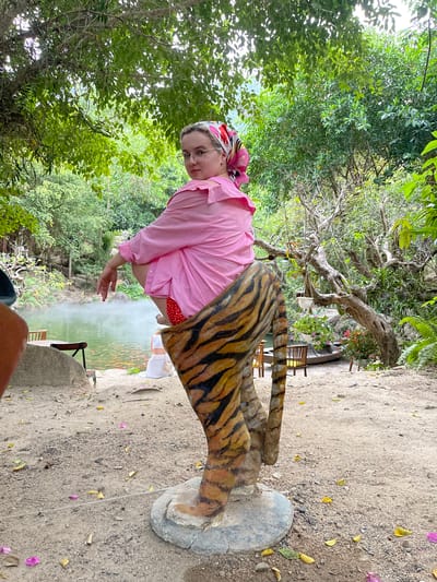 An outdoor scene in Nam Nha Trang, Vietnam, features a person posing playfully amidst lush greenery. A light-skinned individual, likely a woman, is centered in the frame, wearing a bright pink long-sleeved shirt, red polka-dotted shorts, and a colorful headscarf. She wears glasses and looks towards the camera with a neutral expression.
She stands next to a large, decorative tiger-striped prop or statue, with its legs and tail prominently visible in the foreground. Her right hand is extended slightly towards a misty, green-tinted body of water, possibly a pond or hot spring, which shows faint reddish flecks, suggesting fish. In the midground, near the water, several wooden chairs and tables are visible, indicating a seating area.
The ground is a gravelly dirt path, sparsely covered with fallen leaves in shades of yellow, purple, and green. The background is filled with dense, vibrant green foliage and tall trees with substantial trunks and branches, characteristic of a tropical or subtropical environment. The bright, natural lighting indicates it is daytime, and the mist rising from the water suggests warm, possibly humid conditions. The overall impression is one of a relaxed, leisure activity in a natural or park-like setting. No visible text is present in the image.