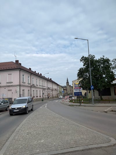 The image shows a street scene in Limanowa, Poland. A light pink building with white trim and numerous windows lines the left side of the street. Across the street, a large tree with dense foliage stands near several buildings, one of which has a sign reading "OPTYK." A silver Volkswagen Caddy van is parked on the right side of the road, facing forward. A cobblestone sidewalk and street are visible in the foreground. The sky is overcast with light clouds. Several cars are present on the road, and two pedestrians are walking on the sidewalk. Construction barriers with red and white stripes are positioned along the side of the road. A church steeple is visible in the distance down the street.
