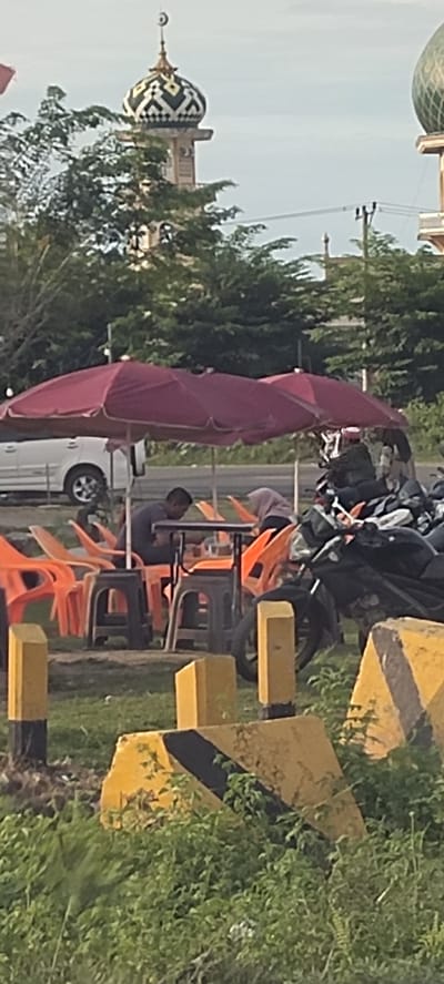 The image presents a scene at what appears to be a roadside eatery in Keupoknibong, Indonesia. Two individuals are seated at a square table under the shade of red umbrellas, engaged in an activity that appears to be eating. The seating consists of orange plastic chairs and dark stools. Motorcycles are parked nearby. The location seems to be adjacent to a road, with a white van parked in the background. In the far background, structures resembling mosques are visible, characterized by distinctive domes. The foreground features yellow and black striped road barriers, partially obscured by vegetation.