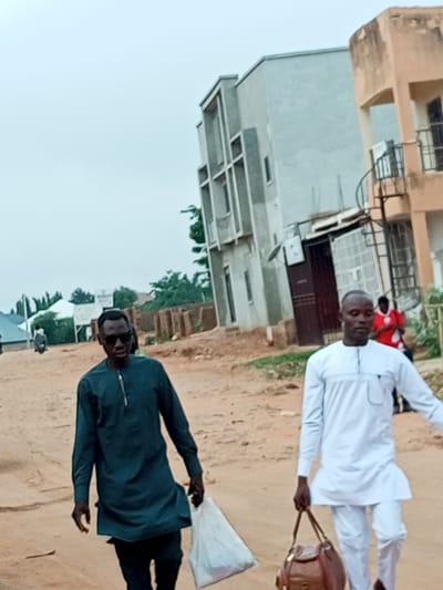 In Wuro Dole, Nigeria, two men are walking on a dusty street. The man on the left wears a dark green, long-sleeved tunic and dark pants, and carries a white plastic bag in his right hand. He is wearing dark sunglasses. The man on the right wears a white, long-sleeved tunic and white pants, and carries a brown leather bag in his left hand. In the background, there are unfinished buildings, a building with a light peach colored facade, and some trees. Other people and a motorcycle are visible in the distance. The sky is overcast.