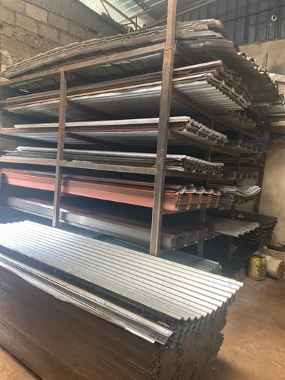 The image shows a collection of corrugated metal sheets stored in a metal rack inside a building. The sheets appear to be of varying colors and finishes, including galvanized and possibly painted. The building appears to be a warehouse or storage facility. The specific location is Biashara ward, Kenya. There are no people visible in the image. Other objects include a concrete wall and metal pipes.