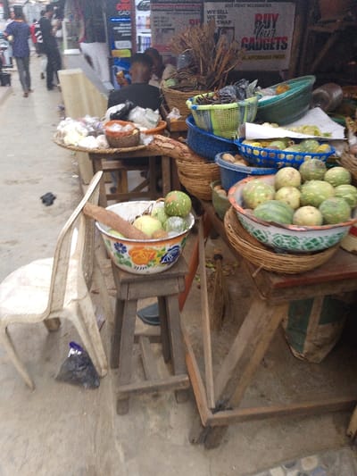 A bustling marketplace scene unfolds with vendors displaying their goods. A variety of fruits and vegetables are arranged in colorful bowls and baskets, perched atop wooden tables and stands. A white plastic chair sits adjacent to one of the stands. In the background, people stroll along the sidewalk, while signs advertise gadgets and accessories.