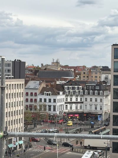 This is an elevated view overlooking a street in Saint-Gilles - Sint-Gillis, Belgium. The scene features a mix of residential and commercial buildings, with architectural styles varying from older brick structures to modern high-rise buildings. A bustling street runs between the buildings, with vehicular and pedestrian traffic evident. Several cars are visible along with pedestrians walking on the sidewalks. Signage and advertisements can be seen on building facades along the street. Metal scaffolding structures are present in the foreground. The sky is overcast with grey clouds, suggesting cloudy weather conditions.