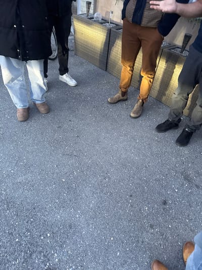 The scene is an outdoor, ground-level view located in Lalden, Switzerland, depicting the lower extremities of at least five individuals standing on a dark grey, granular asphalt surface. Low-angle sunlight from the left casts distinct shadows of the individuals and objects towards the right.

From left to right, the visible attire of the individuals includes:
1.  Light blue, possibly distressed, wide-leg jeans and brown, thick-soled shoes or boots. A dark jacket partially covers the upper body.
2.  Black pants and white low-top athletic shoes. A silver-rimmed bicycle wheel with a dark tire is partially visible behind this individual's left leg.
3.  Brown chinos and brown Chelsea boots. The individual wears a dark top with a brown and dark patterned vest or jacket. The right hand is visible in the upper right quadrant of the figure, displaying the index and middle fingers extended upwards.
4.  Dark grey, possibly distressed, pants and black athletic-style sneakers.
5.  At the bottom right frame edge, the feet and lower calves are visible, clad in light blue jeans and brown leather boots.

In the background, positioned against a light-colored building facade, are two dark grey, rectangular woven-texture outdoor planter boxes or furniture units. These units contain decorative elements, including at least two silver spherical objects and multiple vertical stake-like light fixtures or water features. One feature appears to be a small water feature with a stone or crystal component. The individuals are standing in a static configuration; no overt interactions are discernible beyond their proximity and the single hand gesture.