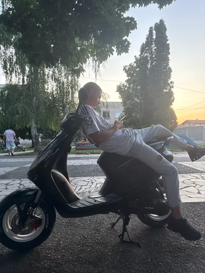 A young person is lying on a black scooter, resting their head on their arm and looking at their phone. They are wearing ripped light-wash jeans, a white t-shirt, and dark sneakers. The scooter is parked on a patterned tiled area next to asphalt. In the background, there are trees, a building, and power lines. The sky is bright with the sun setting, casting a warm glow. The scene appears to be outdoors in a residential area. The overall mood seems relaxed and casual. Text visible on the scooter tire reads "TIMSUN".