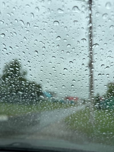 This is a photograph taken through a rain-streaked windshield, looking out onto a street scene. The foreground is dominated by numerous water droplets of varying sizes, distorting the view of the background. Through the water, a blurred landscape is visible, including green grass on the sides of the road, a grey pavement or road surface, and some indistinct objects that appear to be parked vehicles in various colors, including red and green. A utility pole with wires is also visible through the rain. The sky appears overcast. There are no discernible people or interactions. The location is not specified.