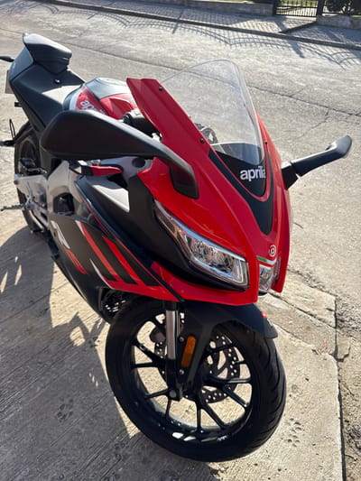 A red and black Aprilia sport motorcycle is parked on a cracked concrete surface, illuminated by bright sunlight. The motorcycle, viewed from a high angle towards its front, features a red front fairing with the "Aprilia" logo on its clear windshield. It is equipped with black handlebars, black mirrors, and prominent LED headlights. The side fairing displays black panels with red and black graphic patterns. The front wheel is black with multiple spokes, featuring a visible disc brake system and an orange reflector on the front fork. Strong, distinct shadows are cast on the ground from the motorcycle and a dark metal fence visible in the upper background. The scene depicts a stationary vehicle in the city of Paisievo, Bulgaria. No individuals, actions, or interactions are present.