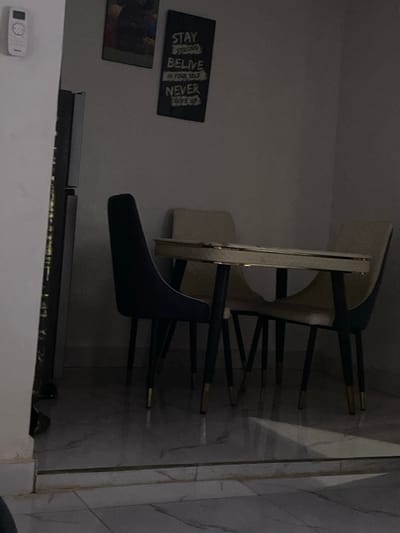 The image depicts a dimly lit indoor residential space, identified as a location in Okpanam, Nigeria. The viewpoint is from a slightly elevated position, looking into a room with light-colored, veined tile flooring. A raised threshold separates the foreground from the main area.
In the center of the frame, a dining set is visible. This includes a rectangular table with a light-colored surface, possibly cream or white, featuring a metallic gold or brass edge trim, supported by four dark-colored legs with gold-tipped feet. Surrounding the table are four chairs, three of which are clearly discernible. On the left, a dark blue or green velvet-upholstered chair with a high back is positioned. On the right, two lighter-colored, possibly cream or beige, upholstered chairs with similar dark legs and gold-tipped feet are partially visible behind and beside the table.
Against the rear wall, two framed items are mounted. The left frame contains a dark-toned image, possibly an abstract or stylized figure. The right frame is a dark, rectangular sign or poster with white text that reads: "STAY STRONG BELIEVE IN YOURSELF NEVER GIVE UP". To the far left of the image, partially obscured by a wall or door frame, a tall, dark, reflective appliance, consistent with a refrigerator, stands. Above and to the left of this appliance, a white, rectangular remote control unit is mounted on the wall. No individuals are present in the scene, and no actions or interactions are depicted.