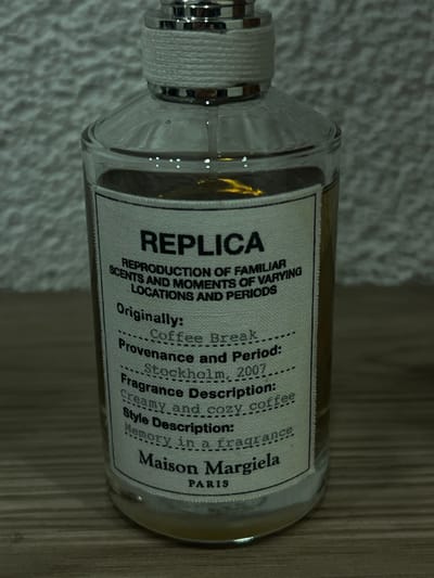 A clear glass perfume bottle with a silver cap and white twine is displayed on a neutral surface. The label reads "REPLICA" in bold, followed by "REPRODUCTION OF FAMILIAR SCENTS AND MOMENTS OF VARYING LOCATIONS AND PERIODS." Details include "Originally: Coffee Break," "Provenance and Period: Stockholm, 2007," and "Fragrance Description: Creamy and cozy coffee." The label finishes with "Style Description: Memory in a fragrance" and "Maison Margiela PARIS."