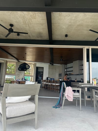 The image shows a partially covered patio with a concrete ceiling and wooden beams. The patio is located in Santa Fe, Costa Rica, with coordinates 9.647° N, 85.166° W. There are two wooden chairs with grey cushions, one with a throw blanket draped over it, and another with a pillow on the seat. There is a large grey, upholstered chair in the foreground, and a table with a wooden chair. The patio opens to a spacious kitchen with stainless steel appliances and a large window overlooking the outdoors. A man is standing at the kitchen sink, washing dishes. The kitchen is well-lit with recessed lighting. The overall environment is clean and modern.