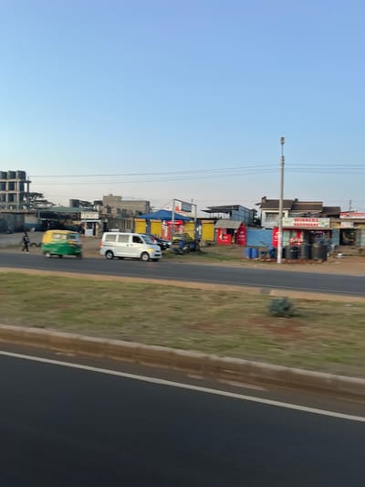 The image depicts a roadside scene in Biashara ward, Kenya, under a clear, light blue sky. In the foreground, a multi-lane asphalt road is visible, separated by a grassy median strip. A white passenger van is moving from right to left on the road, alongside a green and yellow three-wheeled auto-rickshaw (tuk-tuk) traveling in the same direction. A dark-clothed individual walks on the dirt shoulder behind the auto-rickshaw, moving from left to right.

The background features a mix of commercial and unfinished structures. On the far left, a multi-story concrete building with exposed rebar indicates ongoing construction. Towards the right, various small commercial establishments are present, including one clearly marked "WINNERS HARDWARE" with red lettering on a white sign. Outside this hardware store, several black plastic barrels are stacked. Other colorful kiosks, some with red and yellow panels, line the street. Overhead power lines span the scene, supported by utility poles, one of which is prominently positioned on the right side of the road. Sparse vegetation and dirt patches are visible along the roadside and between structures. The overall scene suggests a bustling urban or semi-urban commercial area.