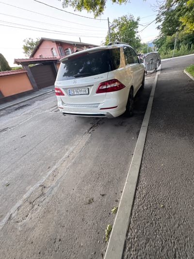 The image, correctly oriented, displays the rear view of a white Mercedes-Benz GL350 SUV parked on the side of an asphalt road in a residential area. The vehicle's license plate, "CB 9699 HK," featuring a "BG" country oval, indicates its registration in Bulgaria, consistent with the given location of Yana.

The scene is set on a clear, sunny day, likely during summer or late spring, given the bright daylight and lush green foliage. The car is parked alongside a concrete curb, which separates the road from a narrow, unpaved strip and a line of verdant trees. In the mid-ground, a large, grey, industrial-style dumpster is partially visible near the curb, nestled among the trees. The background reveals a distant, hazy landscape of hills or mountains under a bright sky.

Behind the parked car, a residential house with a reddish-orange facade, several windows, and a dark-tiled roof is visible. Overhead, power lines stretch across the frame. The asphalt road shows some signs of wear and minor cracks, with small debris scattered near the curb. Visible text on the car includes the Mercedes-Benz logo, "GL 350," and "BLUETEC" on the tailgate, in addition to the license plate details. No people or discernible activity are present, conveying a quiet, everyday street scene.