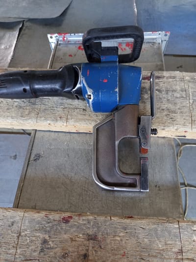 The image shows a close-up, overhead view of a manual metalworking tool, possibly a nibbler or punch, resting on a wooden surface. The tool has a blue and black housing with a black handle. Its metallic lower section appears to be designed for cutting or shaping metal sheets. The tool is positioned horizontally on a piece of weathered wood, which is itself resting on a larger, grey surface. In the background, glimpses of other metallic and possibly fabric materials are visible, suggesting a workshop or industrial setting. The overall impression is one of a functional, potentially used tool in an area dedicated to metal fabrication. The location in Selyatino, Russia is not visually discernible from the image itself.
