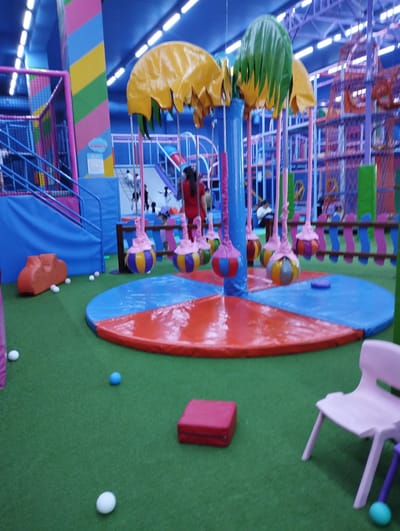 The image depicts an indoor children's play area, likely a playground or amusement center, situated in Talas City, Kyrgyzstan. The environment is vibrant and colorful, with an artificial turf floor. Prominently featured is a circular carousel-like structure with hanging, colorful orb-like decorations and large, umbrella-shaped canopies in yellow and green. In the foreground, there are several white and one blue ball scattered on the green floor. To the left, an orange, cloud-shaped cushioned seat is visible, along with white balls. A pink plastic chair and a red cushioned block are in the right foreground. In the background, various play structures can be seen, including slides, climbing frames, and soft barriers, with some individuals, presumably children and adults, present within the play area. The overall impression is one of a recreational facility designed for children's entertainment.