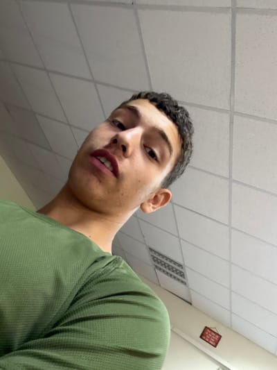 The image displays a young male individual captured from a low-angle perspective, looking downwards towards the camera. He possesses short, dark, curly hair and fair-to-olive skin, with his mouth slightly ajar, revealing upper teeth. The subject is attired in a long-sleeved, olive-green shirt with a visible texture. The background depicts an indoor setting with a white suspended acoustical tile ceiling, featuring integrated rectangular light fixtures or air vents. A light-colored wall is visible on the right side of the frame, bearing a small, rectangular red sign with a decorative border and partially obscured text, consistent with Hebrew script, hanging near the ceiling. This scene is located in Ness Ziona, Israel.