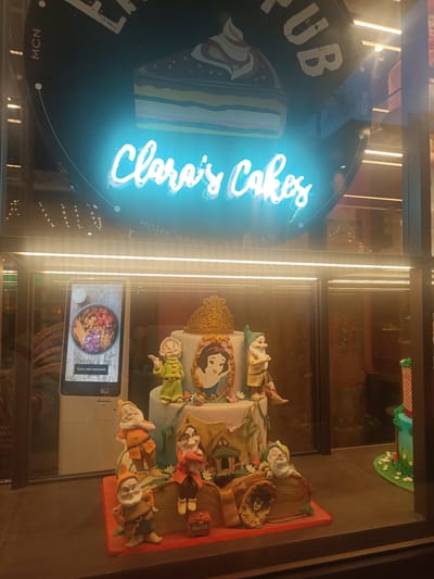 The image shows a storefront display window for "Clara's Cakes" in San Giovanni la Punta, Italy. A large, illuminated sign with the words "Clara's Cakes" in neon blue is displayed prominently. Below the sign, a detailed cake is showcased, decorated with figures of Snow White and seven dwarfs, as well as a miniature house and a tree. To the left of the cake, a digital screen displays food items and ordering options. The display is illuminated by bright lights.