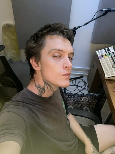 An indoor, horizontally-oriented image (rotated 90 degrees counter-clockwise) depicts a young individual, likely male, with short, dark, curly hair and light skin. The person is wearing a dark grey t-shirt and has a distinctive tribal-style tattoo on their neck. Their eyes are partially closed, and they are looking upwards and slightly away from the camera, giving a contemplative or relaxed impression.
The setting appears to be a home music studio or creative workspace. To the left of the subject's head, a black microphone stand is visible, extending upwards towards what looks like dark grey acoustic paneling on the wall. In the background, behind the person's head, a wooden desk holds a white electronic music production device or synthesizer with colorful knobs and buttons, bearing the visible brand name "ARTURIA." Below the desk, a patterned rug with intricate designs covers the floor, with loose cables coiled on it. A dark-colored office chair is partially visible under the desk, and a bare leg with tattoos and an arm are seen on the right side of the frame. The lighting is ambient, suggesting an indoor environment. The scene captures a moment of rest or introspection within a creative space.