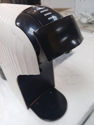A white and black coffee maker sits on a white countertop in Rezina, Moldova. The coffee maker has a ribbed white body and a black top with several horizontal lines. A black plastic piece extends from the top of the coffee maker. The countertop has a faint circular pattern.