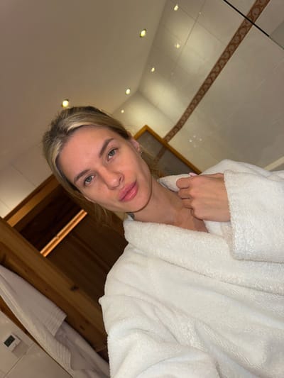 A woman is featured in a close-up, rotated portrait, appearing to be in an indoor setting. She has light hair, fair skin, and light-colored eyes, with her lips slightly parted, creating a neutral and calm expression. She is wearing a white, fluffy bathrobe, and her left hand is visible, gently holding the fabric near her neck. A subtle tattoo is partially visible on her lower neck or shoulder area.
The background indicates a well-lit interior, likely a bathroom or spa. The ceiling is light-colored, featuring several recessed spotlights that provide illumination. A decorative border with a brown, patterned design runs along what appears to be a wall or a different section of the ceiling. In the lower right quadrant of the image, a distinct wooden structure is visible, suggesting wooden paneling or possibly part of a sauna. Below this, a white towel is draped over an unseen object, and a small, partially obscured digital display (possibly a thermostat or clock) can be seen.
The activity appears to be a relaxed moment, potentially a selfie or a photograph taken after a shower or spa treatment. The artificial lighting points to an indoor scene, not indicating a specific time of day. Given the context of Going am Wilden Kaiser, Austria, the environment aligns with a hotel or resort spa. No discernible text is clearly visible in the image.
