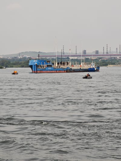 A blue and red barge is on the water with a factory in the background under a cloudy sky. Two inflatable boats are near the barge. One boat has three people in it, all wearing orange life jackets. The other boat has one person wearing a blue shirt and an orange life jacket. The water is choppy with small waves. The barge has Russian writing on its side. In the background, there is a bridge with a red and blue stripe, and a line of industrial chimneys. The city of Pesčanka, Russia, is visible on the shore.