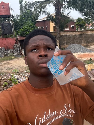A young man is holding a sachet of "miriugo" sachet water up to his lips, seemingly about to drink from it. He is wearing a brown long-sleeved shirt with white text. The photo appears to have been taken outside, in what looks like an urban area or a roadside in Orifite, Nigeria, indicated by the sachet water brand. In the background, there are buildings, palm trees, and some piles of rubble or construction materials. There's also what looks like a water tank. Litter is scattered on the ground near the subject.
