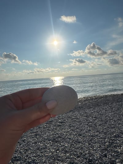 A hand holds a smooth, light-colored stone against a backdrop of the sea and sky. The sun shines brightly in the upper left of the image, casting a glare on the water and creating lens flare. The sky is a clear blue with scattered white clouds. The sea is calm, with gentle waves lapping at a shore covered in small, dark pebbles. The overall mood is peaceful and serene, suggesting a moment of quiet contemplation at the beach. The image appears to be taken during the day, likely in the late morning or early afternoon, given the bright sunlight. The location context suggests Alakhadzi, Abkhazia, a coastal town known for its pebble beaches. There is no visible text in the image.