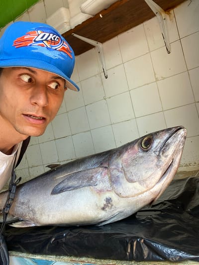 A man wearing a blue baseball cap with an "OKC" logo is posing for a selfie with a large, whole, silvery-grey fish. The fish is upright, positioned to the right of the man, and its head is near the top of the frame. The man's face is tilted upward toward the fish. The background consists of white tiled walls, brackets, and black material (possibly a plastic bag or liner). The scene appears to be indoors, potentially in a market or food preparation area. The photograph was taken in San José de Barlovento, Venezuela.