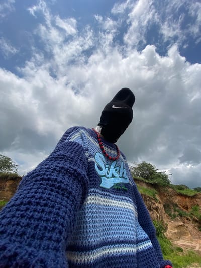 A low-angle shot captures a person from the chest up, facing away from the camera. The person wears a black beanie with a white Nike logo and a black face covering, obscuring their identity. A red and black beaded necklace adorns their neck, resting on a blue, white, and grey striped knitted sweater. The sweater has a graphic design of light blue letters spelling "Sikh" across the chest.
The background is dominated by a dramatic cloudy sky, with patches of blue peeking through. Below the sky, a rugged, earthy cliff face rises, covered in sparse green vegetation. The terrain suggests an outdoor, possibly rural or semi-rural, environment. The lighting indicates it is daytime, with the clouds diffusing the sunlight. The perspective implies the photographer is positioned below the subject, looking upwards. The location is identified as Ufu Esioni, Nigeria.