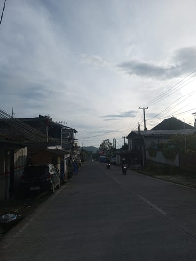 A quiet, paved road stretches into the distance, flanked by buildings and houses on either side. Two motorcycles are seen traveling along the road. A dark-colored car is parked on the left. Above, a cloudy sky dominates the scene, with power lines stretching across the frame. The overall atmosphere suggests a peaceful, early morning or late afternoon in a suburban area.