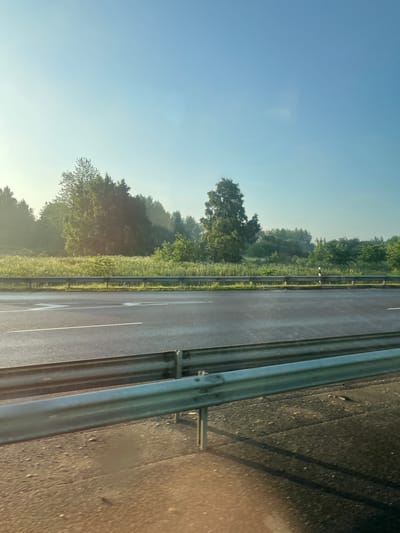 A highway is bordered by a metal guardrail on either side of the scene. The highway has two lanes in each direction. The left side of the highway is lined with trees and other vegetation. The sky is clear and blue. The scene appears to be a highway in Maardu, Estonia.