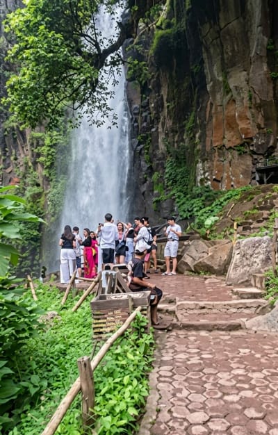 A group of tourists stands near a tall waterfall, posing for pictures and observing the scenery. The waterfall cascades down a rocky cliff face covered in lush green vegetation. A stone pathway winds through the area, leading to a viewing platform. The environment is tropical, with dense foliage and a humid atmosphere. The tourists are casually dressed, suggesting a leisurely outing. The scene captures a moment of enjoyment and exploration in a natural wonder.