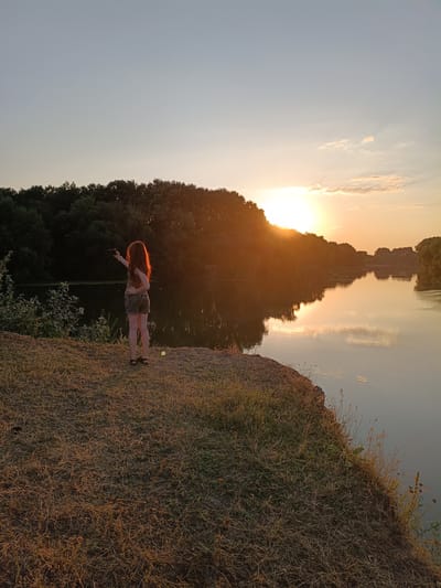 A young woman with long, reddish-brown hair is standing on a grassy embankment by a river at sunset. She is wearing a brown tank top and camouflage shorts, and is holding a smartphone out to take a picture. The sun is setting behind a line of trees on the opposite bank of the river, casting a warm glow over the scene and reflecting on the water. The sky is a pale blue with a few wispy clouds. The embankment is covered in dry grass and some green foliage. The river is calm and appears wide. This scene is consistent with the landscape along the Nistru River near Nezavertaylovka, Moldova.