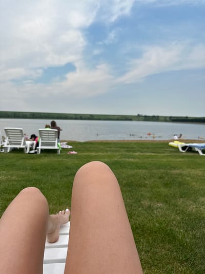 The image depicts a close-up view of a person's legs and feet resting on a white lounge chair. The background shows a body of water, likely a lake, with several people swimming and a treeline in the distance. The sky is partly cloudy. The scene appears to be a recreational area, possibly a beach or park, with lounge chairs set up on a grassy lawn. The location is specified as Emel'yanovo, Russia.