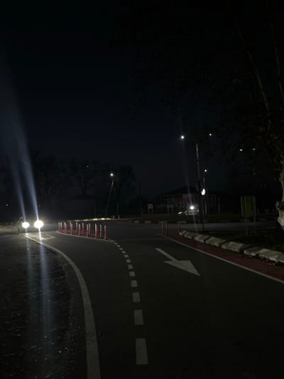 The image displays a nighttime scene of a road junction, specifically a roundabout, located in ლესის თემი, Georgia. The dark asphalt road curves through the foreground, featuring a solid white line along its inner edge and a broken white line separating a lane on the left. This lane is further defined by a series of approximately fifteen orange and white traffic delineator posts. A large white arrow is painted on the asphalt, indicating a right turn. The road's right edge features a raised concrete curb with a red-painted top surface.
On the left, a vehicle approaches with bright headlights, casting prominent, elongated beams of light onto the road surface. A second, lighter-colored vehicle with illuminated headlights is partially visible on the far side of the roundabout. Multiple streetlights provide artificial illumination across the scene, appearing as distinct light sources and glow points. In the background, several dimly lit buildings, including a two-story structure, are visible amidst what appear to be bare deciduous trees. Two red-and-white chevron signs, indicating a left curve, are positioned on the far side of the roundabout. A tree with dark foliage is present on the far right edge of the frame. The overall environment is characterized by low natural light conditions, with visibility primarily maintained by the artificial light sources and vehicle headlights.