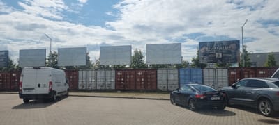 In Mroków, Poland, on a paved lot, a white van is parked facing away from the camera. To its right, a black Audi TT is parked. Further to the right, a grey Audi SUV is parked. Behind the vehicles, a line of shipping containers is visible, topped by several large billboards. The sky above is filled with scattered clouds.