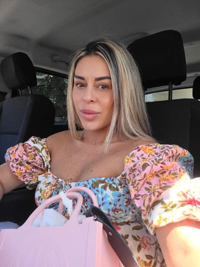 A woman with blonde hair streaks and a floral off-the-shoulder top is inside a car. She is looking at the camera with a slight smile. In front of her, a light pink tote bag is visible, containing white fabric. The interior of the car, with its dark seats and headrests, is apparent, as are trees visible through the car window, suggesting an outdoor setting. The location is the city of Peshtera, Bulgaria.