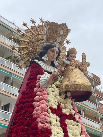 A large, ornate religious statue depicting the Virgin Mary and the Infant Jesus is prominently displayed outdoors in the city of Paiporta, Spain. The statue of the Virgin Mary, with dark hair and a serene expression, wears an elaborate gold crown with radiating spikes, large white ornate earrings, and a ruffled white collar. Her garment is extensively adorned with rows of real flowers in dark red, light pink, and white/cream, possibly carnations, arranged in vertical stripes and forming some patterns. Her right hand delicately holds a small green stem. The Infant Jesus, held by Mary, wears a smaller gold crown, an ornate gold robe with embossed patterns, and a white ruffled collar. He holds a golden cross or scepter upright in both hands. The statue is positioned high, suggesting it is on a float or platform. In the background, out of focus, are multi-story residential buildings with light-colored facades, dark-framed windows, and balconies with metal railings. Some balconies feature plants and air conditioning units; a green awning is visible on one lower balcony to the right. The sky is overcast and grey.