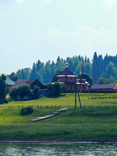 The image depicts a serene summer landscape on the outskirts of Ust-Kem, Russia, likely near a body of water, possibly the Kem River. In the foreground, the rippling surface of the water reflects the light. Beyond the water, a grassy embankment rises, dotted with numerous wooden benches arranged in rows along its crest. A wooden walkway with steps ascends the embankment, leading towards a cluster of buildings.
Dominating the mid-ground is a wooden Orthodox church with a dark brown roof and golden cupolas, its exterior constructed from light-colored logs. Adjacent to the church, to its right, is a smaller wooden structure with a dark roof, possibly a chapel or auxiliary building. Further to the left, an older, dilapidated wooden house with a dark, weathered roof stands partially obscured by lush green foliage. A tall wooden utility pole stands prominently in front of the church.
The background is characterized by a dense forest of coniferous trees, their dark green canopies creating a stark contrast against the pale blue sky, which is sparsely populated with white clouds. The overall impression is one of a peaceful rural setting, bathed in the soft light of a summer day. There are no people visible in the image.
