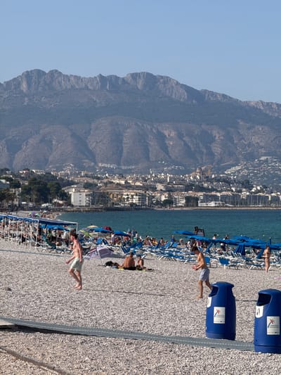 The image depicts a crowded beach scene in Pla Parcial Platja, Spain, with a vast expanse of mountains forming the backdrop. The beach is covered in small, light-colored pebbles, and numerous lounge chairs with blue umbrellas are set up. Several people are visible on the beach, some sunbathing, others walking, and a few playing with a ball. The sea is a vibrant turquoise. In the foreground, two blue trash bins are positioned. The city, with its white buildings, is nestled at the foot of the mountains, extending along the coastline. The sky is clear and blue.