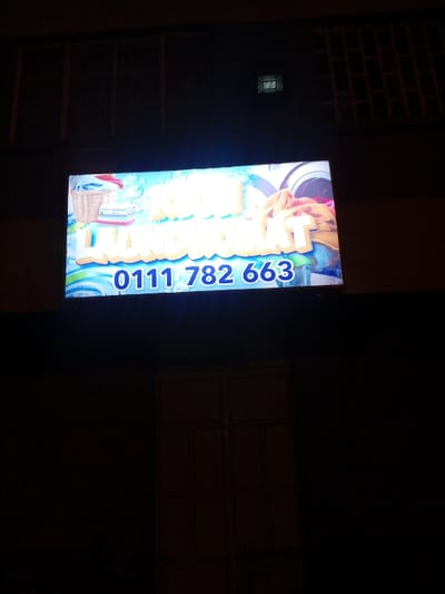 A brightly illuminated sign displays the name "Laundromat" in large, vibrant white letters against a turquoise backdrop featuring animated water swirls and laundry-related imagery. A phone number, "011 782 663," is prominently displayed vertically on the right side of the sign in blue. The sign is mounted on a dark wall adjacent to a brick section and a multi-paneled window. The surrounding area is dimly lit, highlighting the sign as the focal point.
