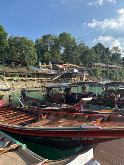 The image depicts a scene in Ban Ta Khun, Thailand. Foreground features show several traditional Thai longtail boats moored alongside a wooden dock. The boats are painted in earthy tones of brown and red, with some displaying intricate scripts and numbers. In the middle ground, a multi-tiered concrete structure with staircases ascends towards buildings nestled amongst dense green trees. A sign displaying "KHAOSOK NATIONAL PARK" is visible. The background consists of a clear blue sky with scattered white clouds.