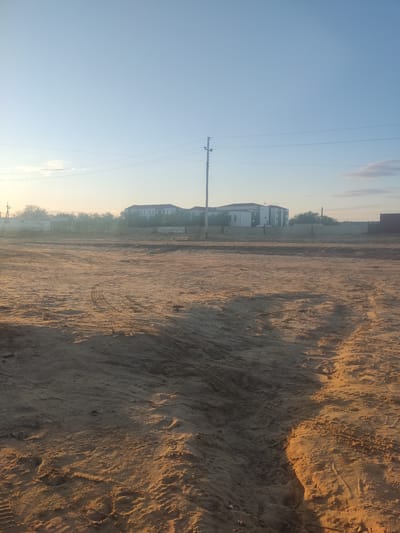 This image shows a rural landscape with a sandy ground in the foreground, marked with tire tracks and footprints. In the background, several modern buildings, likely residential or administrative, are visible, surrounded by some trees and vegetation. Power lines and a utility pole are also present, indicating infrastructure in the area. The sky is clear and light blue, suggesting it is either early morning or late afternoon, with the sun casting long shadows on the ground. The scene appears to be located on the outskirts of Aral, Kazakhstan, a city known for its proximity to the drying Aral Sea.