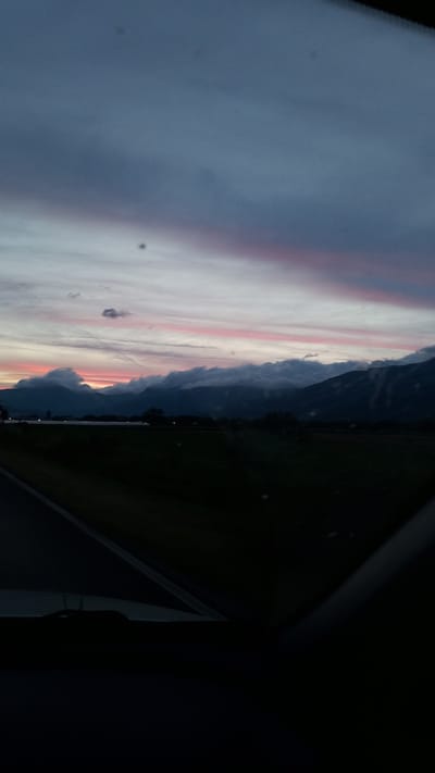 The image captures a twilight scene with a dramatic sky over a landscape. In the foreground, a dark road curves to the left, with a hint of a car's interior visible along the bottom edge. The middle ground is dominated by a dark, silhouetted field of grass. Beyond the field, a range of mountains rises, partially obscured by heavy, dark clouds that are illuminated from below by the fading sunlight. The sky above the mountains displays streaks of pink and orange, transitioning into a deep blue at the top of the frame. A few small, dark spots are visible on the sky. The overall impression is one of a serene, natural landscape at dusk, with the mountains of Gabarevo, Bulgaria, providing a majestic backdrop.