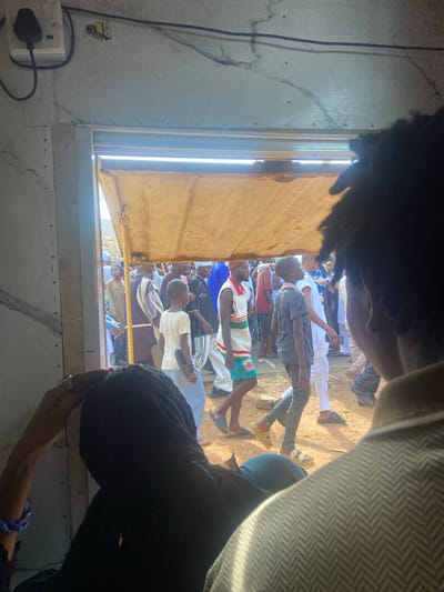 The image depicts a view from inside a building, looking out a window or opening, at a group of people gathered outside. The location is Okengwe - Obehira, Nigeria.
Several individuals are visible outside, many standing or sitting on the ground. Some are wearing casual clothing, including jerseys and T-shirts. Some are barefoot.
The foreground contains elements of the building's interior, including a power outlet, wall, and framing around the opening. There is also a dark shape obscuring part of the view, possibly hair or clothing. An arm and hand are visible in the bottom right corner.