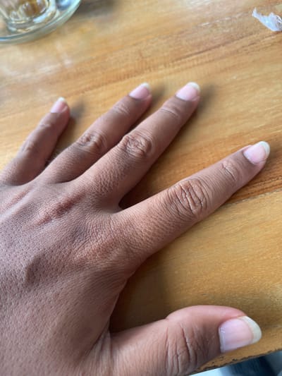The image shows a close-up of a person's hand resting on a wooden surface. The hand is positioned diagonally across the frame, with the fingers spread slightly. The skin tone appears to be medium to dark, and the nails are neatly trimmed. Visible on the hand are the natural creases and texture of the skin, particularly on the knuckles and the back of the hand.
The surface beneath the hand is a wooden table with a visible grain pattern, suggesting it is likely indoors. The lighting appears to be natural, casting subtle shadows and highlighting the textures of the skin and wood. The overall mood is neutral and observational, focusing on the details of the hand and its immediate environment.
In the lower left corner of the image, a portion of a clear glass object, possibly a bowl or cup, is visible, containing a light-colored liquid. The presence of this object might indicate a setting where someone is eating or drinking.
There is no visible text in the image. The scene is simple and focused, presenting a common everyday object and body part in a well-lit, natural setting.