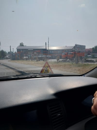 The image depicts a view from inside a vehicle in Belokurikha, Russia. The foreground shows the dashboard and a person's hand with a ring. Outside the windshield, a road is visible, with a construction warning sign featuring a silhouette of a person with a shovel. In the background, a long, modern building with a grey, corrugated roof is situated. Orange trucks are parked in front of the building, and signs indicate "Hotel" and directions to "Автоград" (Avtograd) at a distance of 1.6 km. The environment appears to be a roadside area with some vegetation and a slightly hazy sky.