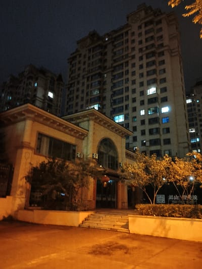 The image captures a nighttime scene in Jiguan District, China, featuring residential buildings and a low-rise structure with an arched entrance. In the foreground, a paved area, likely a street or courtyard, is illuminated by warm-toned artificial light, casting shadows. To the left, a cream-colored building with a decorative cornice and dark windows is visible. Behind it, and dominating the background, are tall apartment buildings with numerous illuminated windows, indicating occupancy. Some trees with golden-hued foliage are present, particularly to the right, suggesting an autumn setting. A sign with Chinese characters and English text, "THE JOY PARK," is partially visible on a low wall near the trees. The sky is dark, indicating nighttime, and the overall atmosphere is subdued, with the lighting from the buildings and street providing the primary illumination. There are no people or overt actions depicted in the scene.