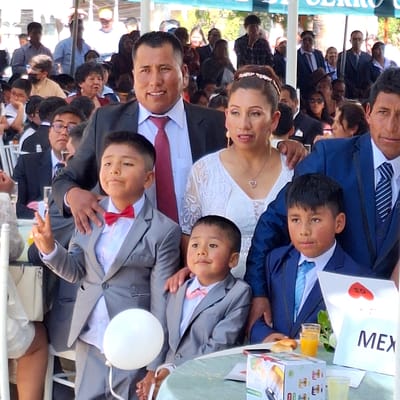 The image depicts a group of people posing for a photo, likely at an event like a wedding, in Cerro Colorado, Peru. At least three generations are visible, and all people seem to be of the same ethnic group. The bride and groom are front and center with what appears to be the groom's sons with them. Most of the males are wearing suits, including boys and groomsmen. There is a white balloon and table with a white 'MEX' sign. More people sit at tables in the background. A 'Cerro Colorado' signpost is vaguely visible.
